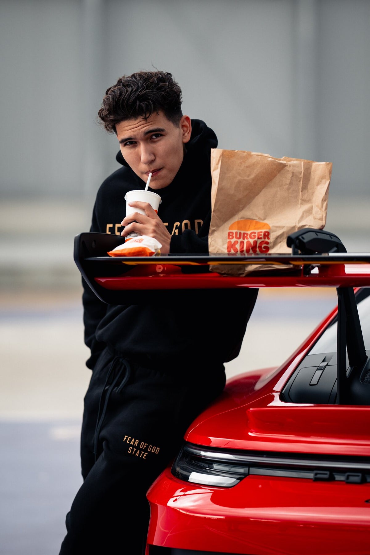 Ádám Péter, founder of OnSize, in a black Fear of God outfit, leaning against a red sports car